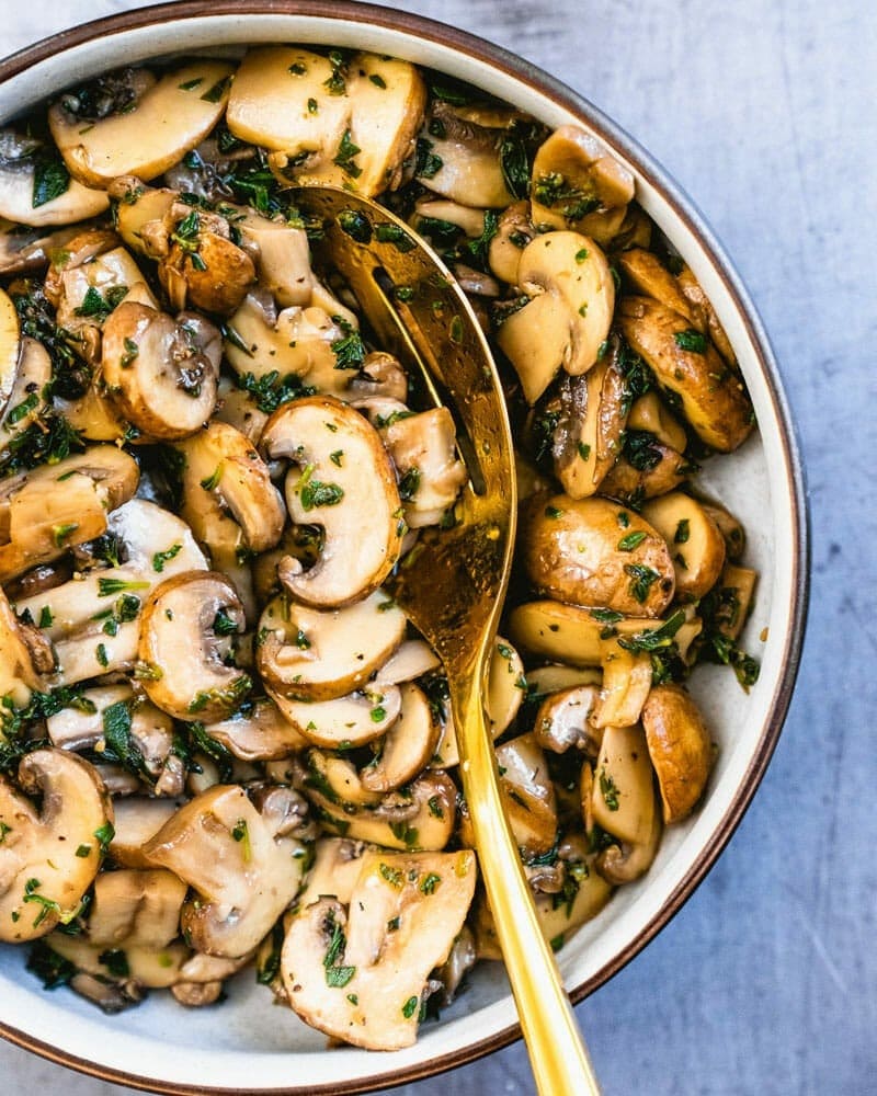 A bowl of sauteed mushroom with herbs tossed by a wooden spoon.