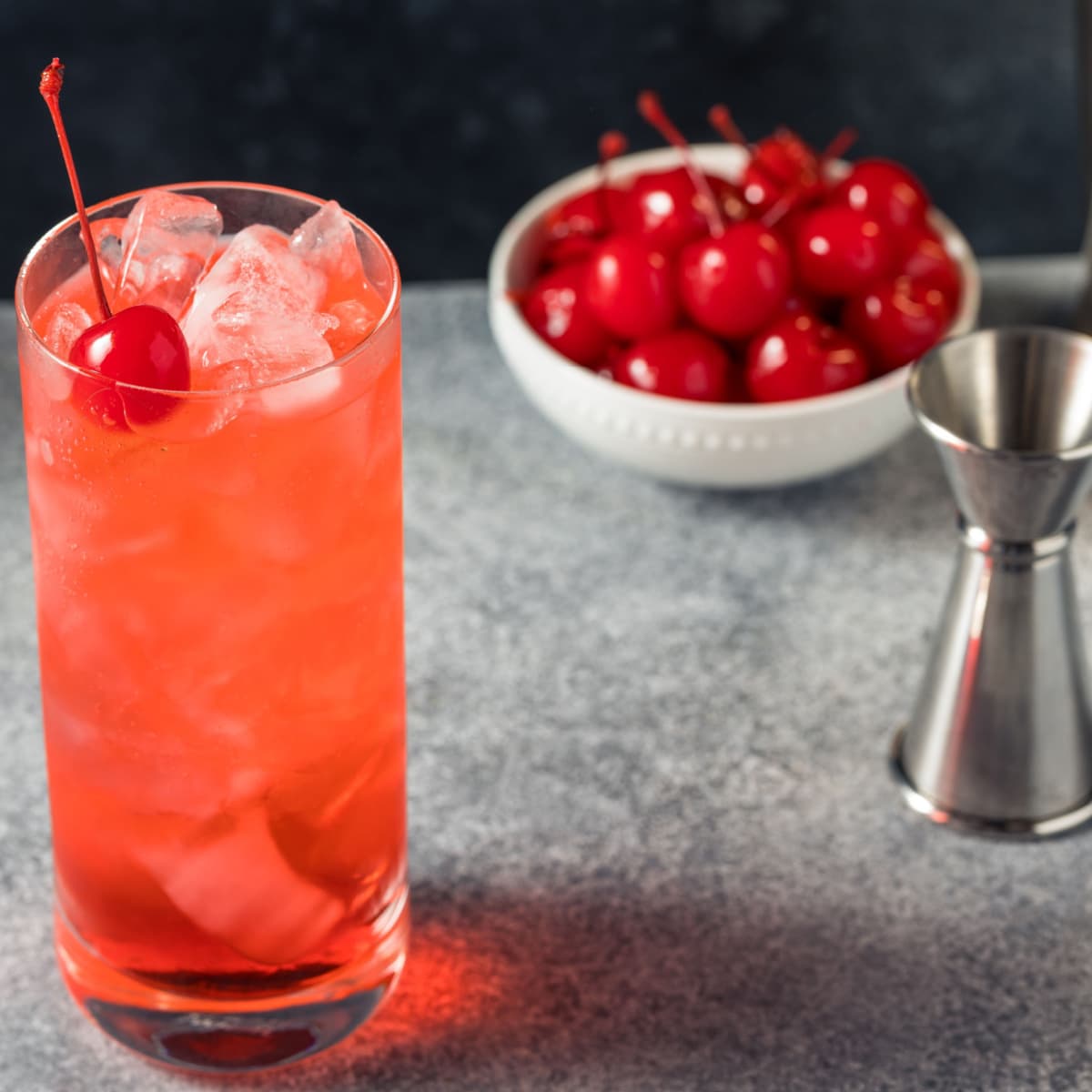 Shirley temple on a tall glass filled with ice beside a bowl of fresh cherries and jigger. 