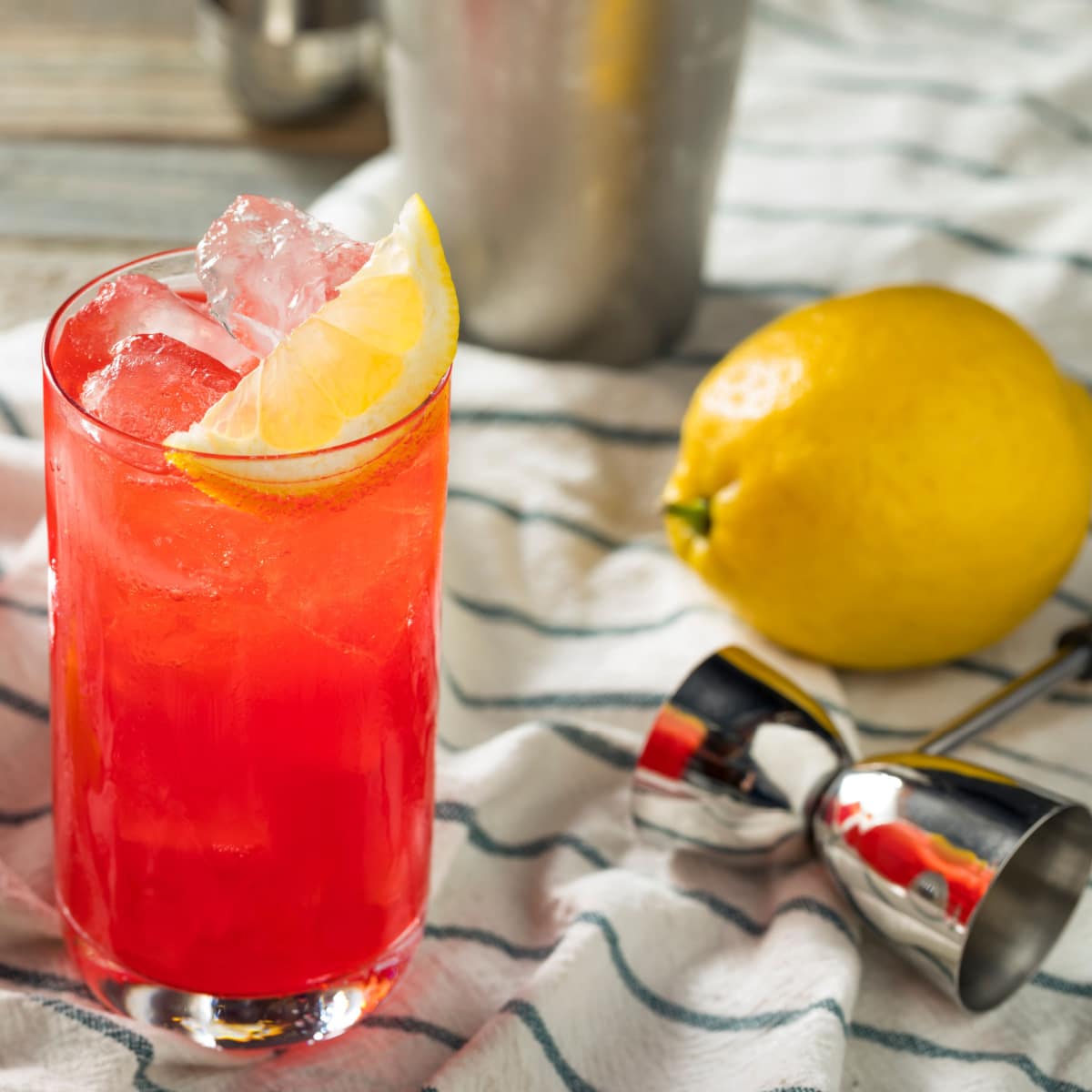 A glass of ice cold red orangey cocktail garnished with lemon slice, whole lemon and jigger beside. 