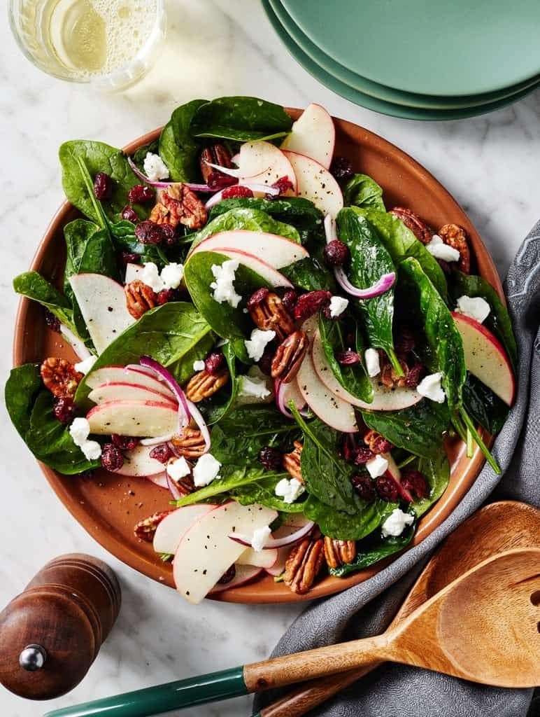 A wooden bowl of spinach salad with crisp apples, dried cranberries, and toasted pecans. 