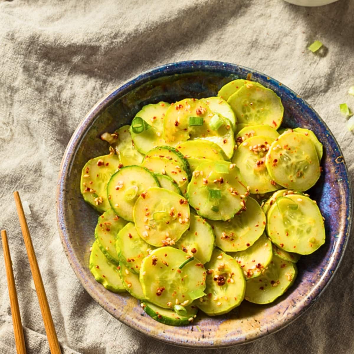 Top view of a bowl of Asian cucumber salad with chili flakes and scallions and a pair of chopsticks beside. 