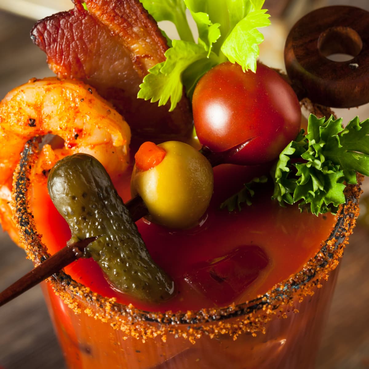 Top view of Tequila Bloody Mary in a glass garnished with shrimp, bacon and olives, pickles and olives on skewers. 