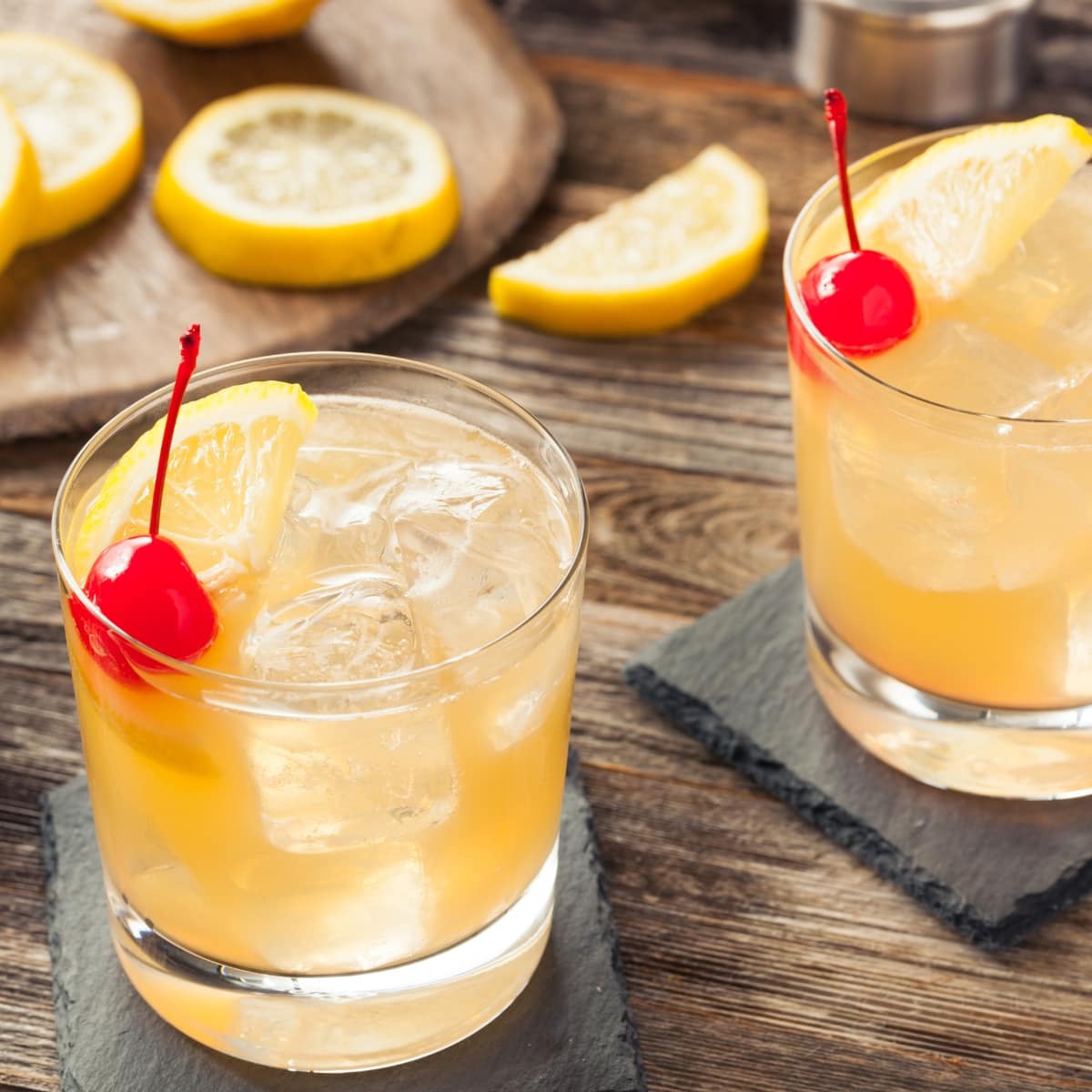 Two glasses of whiskey sour cocktail with ice garnished with cherry and lemon slice. 