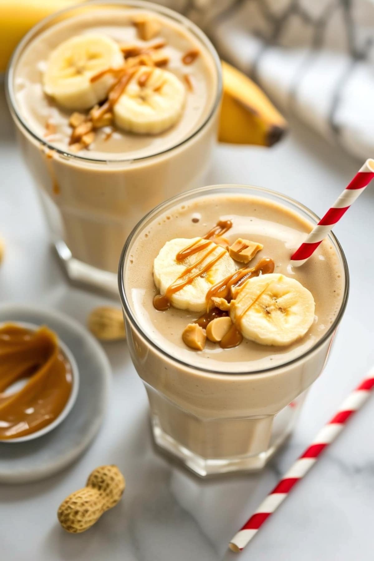 Two glasses of smoothie with peanuts, banana slices, and peanut butter 