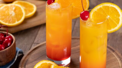 Two glasses of sunrise cocktail on a wooden tray served with orange slices and red cherries garnish.
