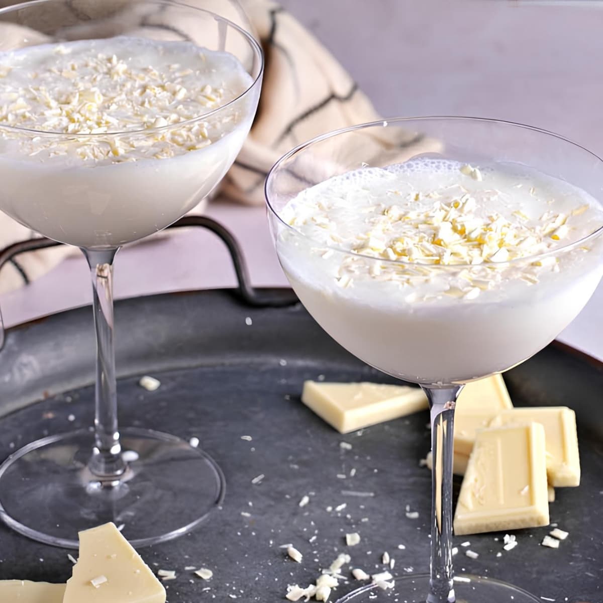 Two servings of white chocolate martini on wine glasses garnished with chocolate shavings on top of a metal tray. 