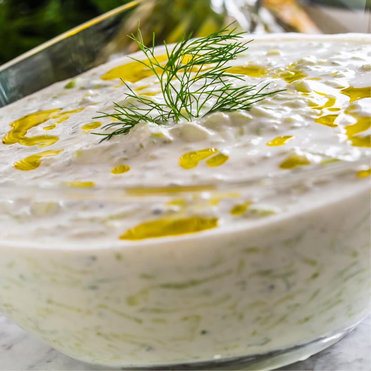 Closeup of tzatziki sauce in a glass bowl with fresh dill leaves garnish and drizzle of olive oil. 