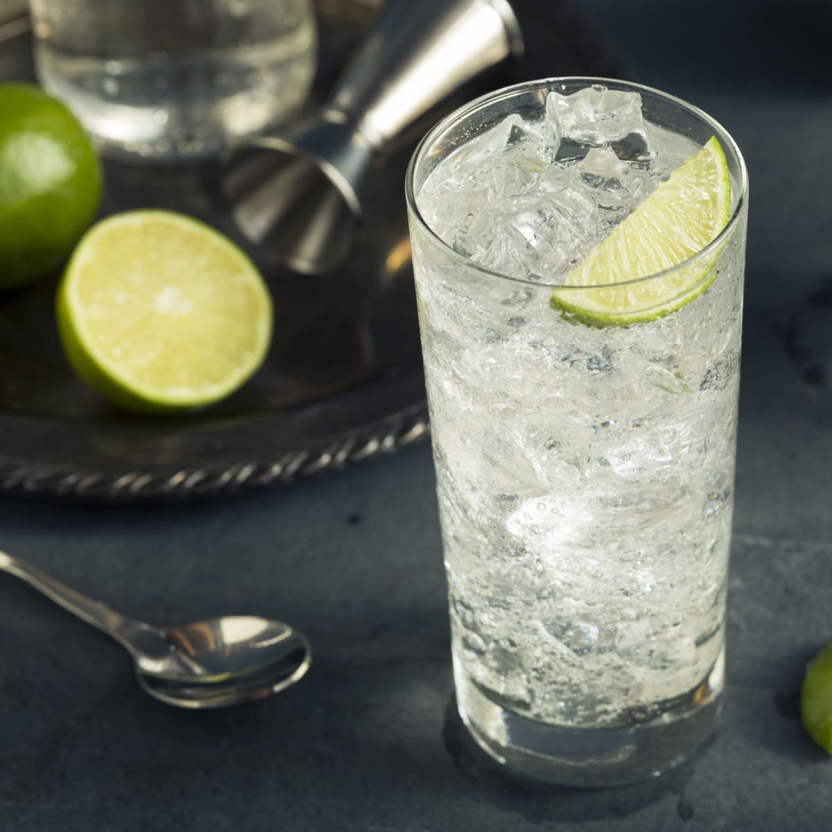  A tall glass of vodka soda filled with ice garnished with lime slice, bottle of vodka, jigger and lemon slice on a steel tray in the background. 