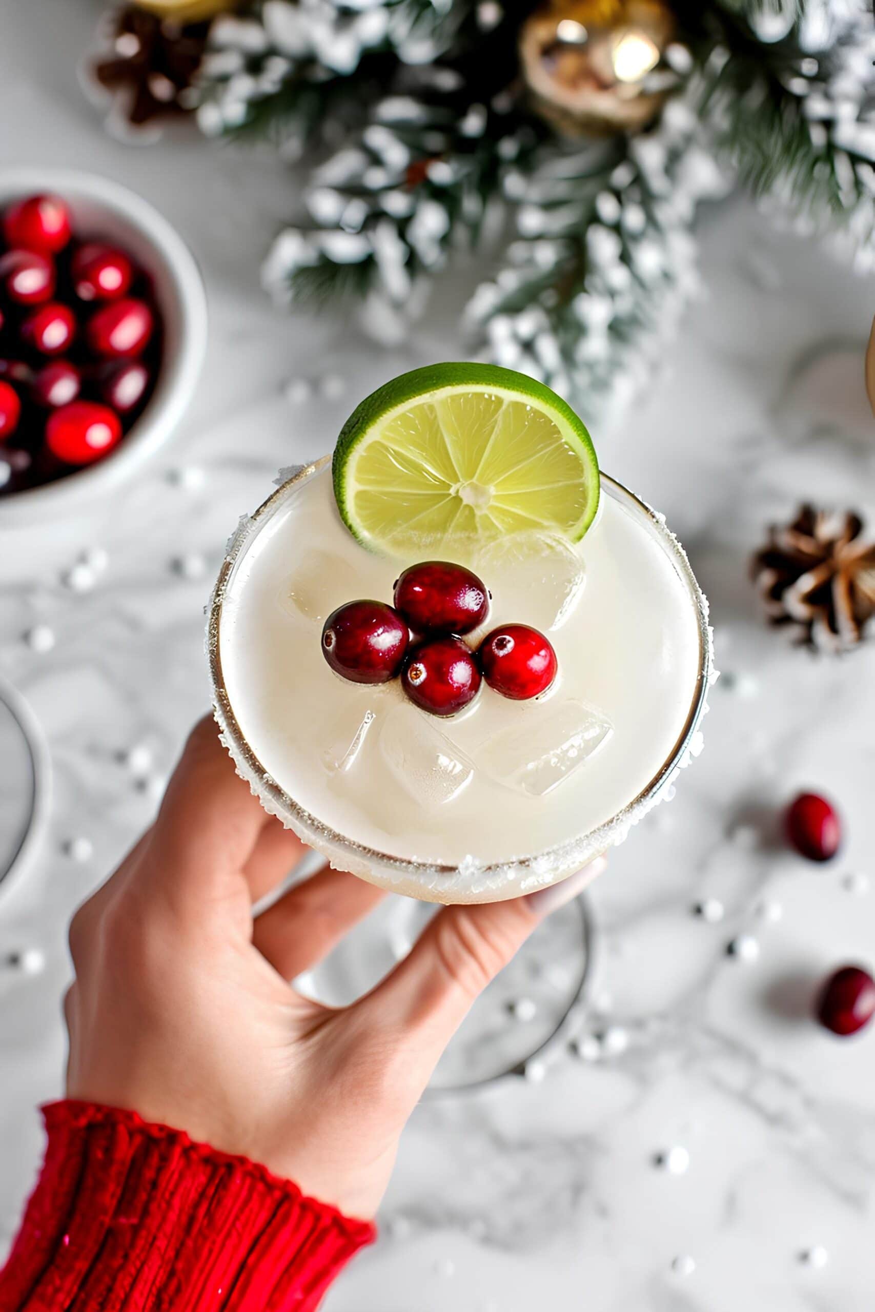 Easy White Christmas Margarita Recipe: A hand holding a white Christmas margarita garnished with lime wedges and cranberries