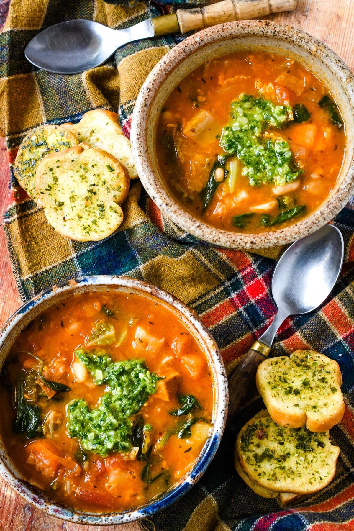 Vegetable soup with vegetables, parsnips, cabbage, spinach, and pesto, served with garlic bread and a spoon on a rustic table cloth