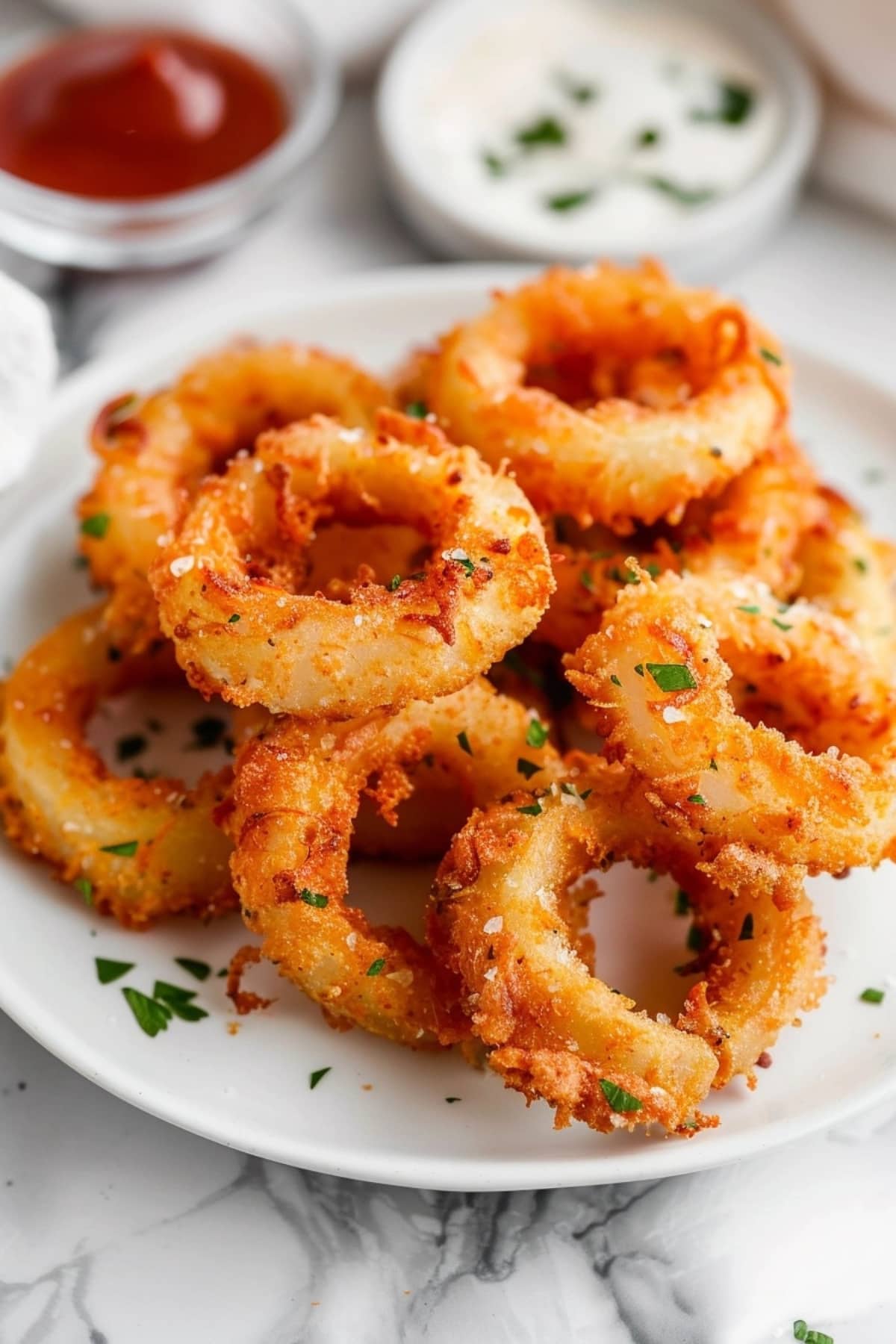 Air fried onion rings in a white plate garnished with chopped herbs and dipping sauce in the background.