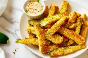 Bunch of breaded air fried zucchini fries in a white plate on a white marble table served with dip.