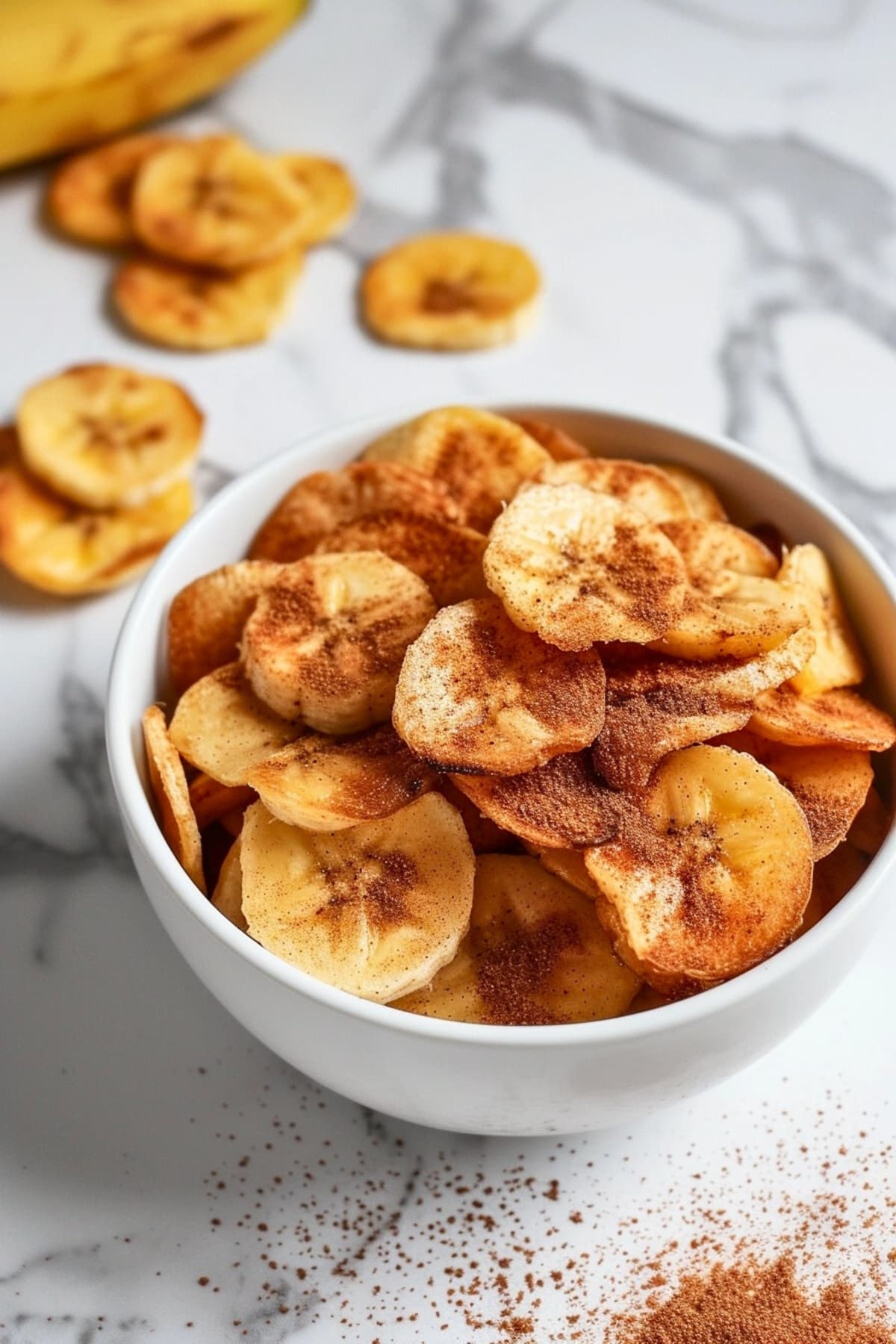 Bowl of banana chips dusted with cinnamon powder in a white bowl.