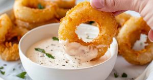Hand dipping air fried onion ring on a bowl of sauce.