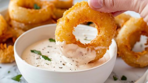 Hand dipping air fried onion ring on a bowl of sauce.