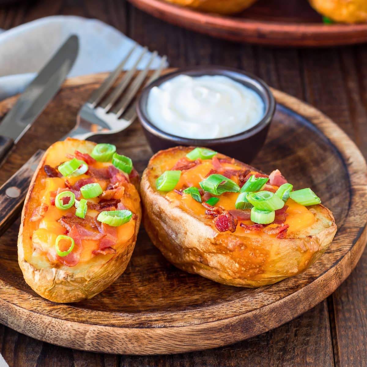 Potato skins with cheesy filling garnished with bacon and chopped onions served on a wooden tray with sour cream.