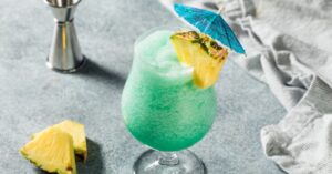 Blue Hawaiian cocktail garnished with pineapple slice and cocktail umbrella on top of concrete table.