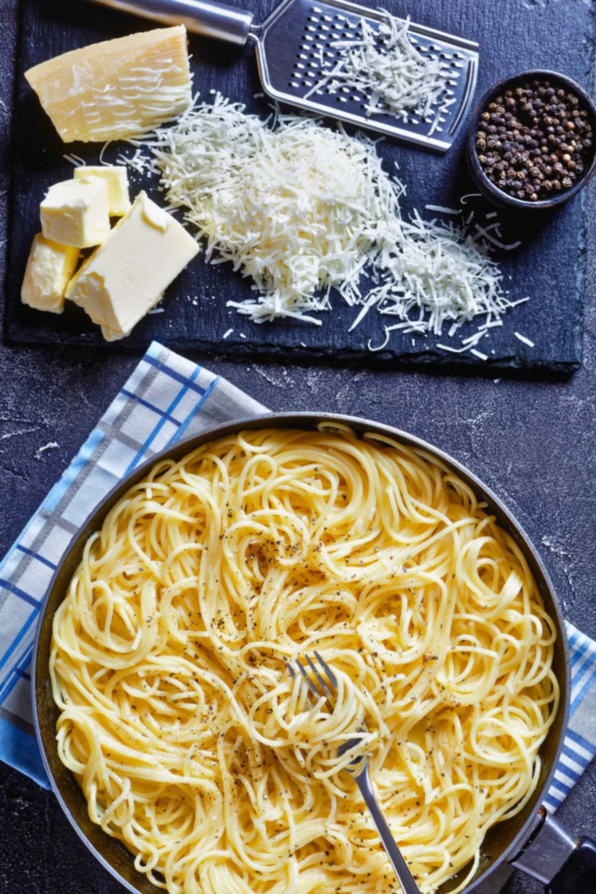 Caco e Pepe in a black pan beside grated cheese ion a black slate.