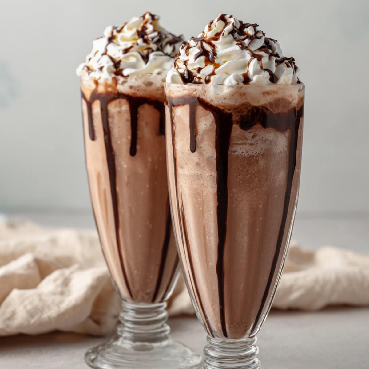 Two chocolate milkshakes with whipped cream in a tall clear glass.
