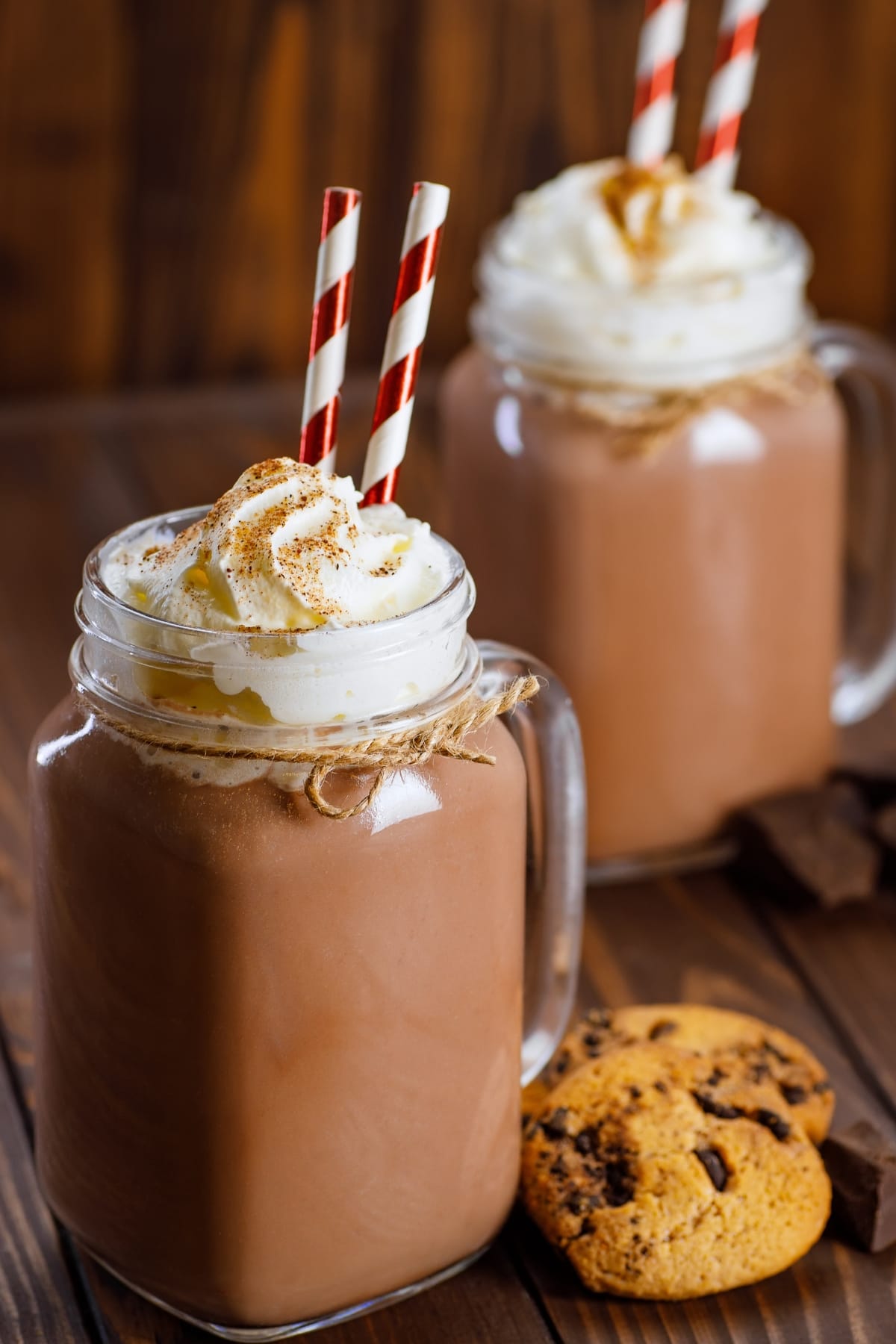 Two mugs of whipped cream topped chocolate milkshake beside cookies and chocolates on a wooden table. 