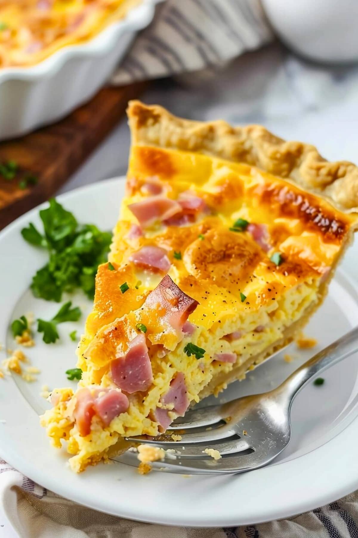 A slice of ham and cheese quiche in a white plate garnished with chopped parsley.