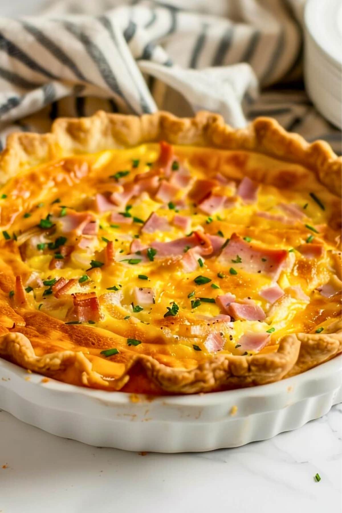 Ham and cheese quiche in a white pie dish, towel beside.