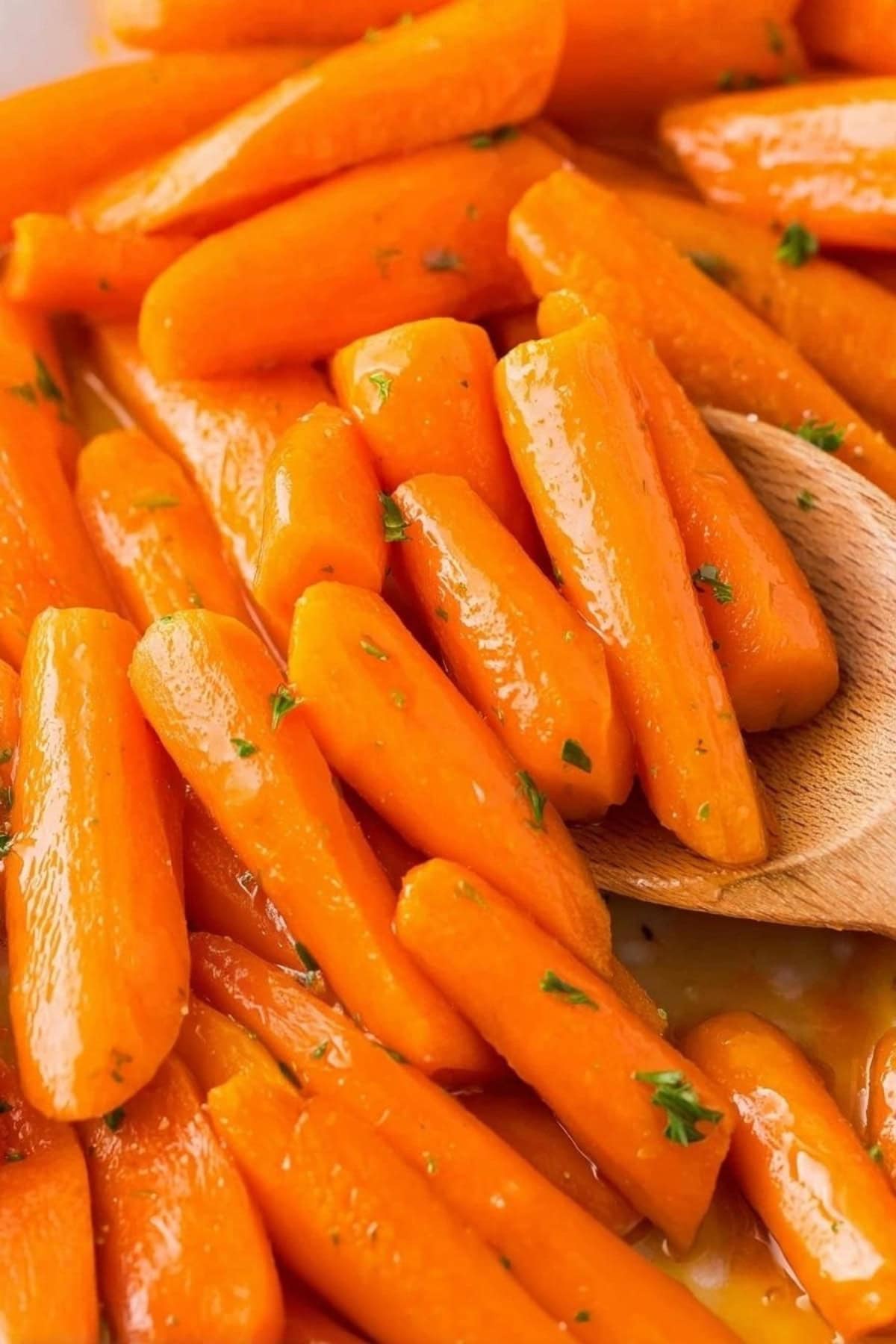 Close-up of glossy honey-glazed carrot pieces garnished with chopped parsley, with a wooden spoon resting beside them.