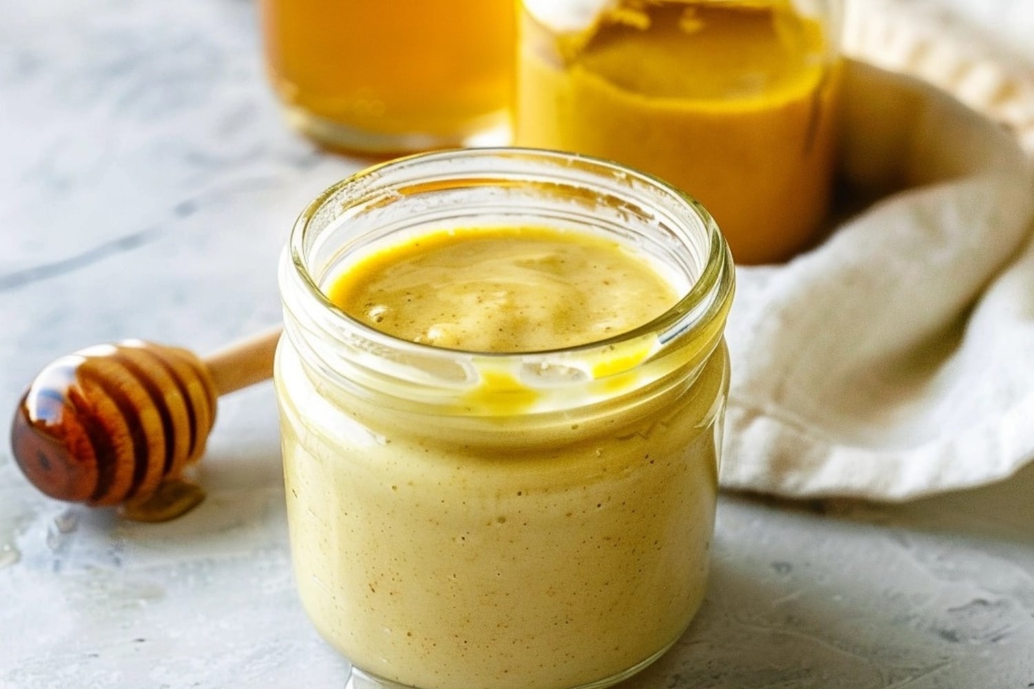 Small glass jar with honey mustard sauce, mustard and honey in jar in the background.
