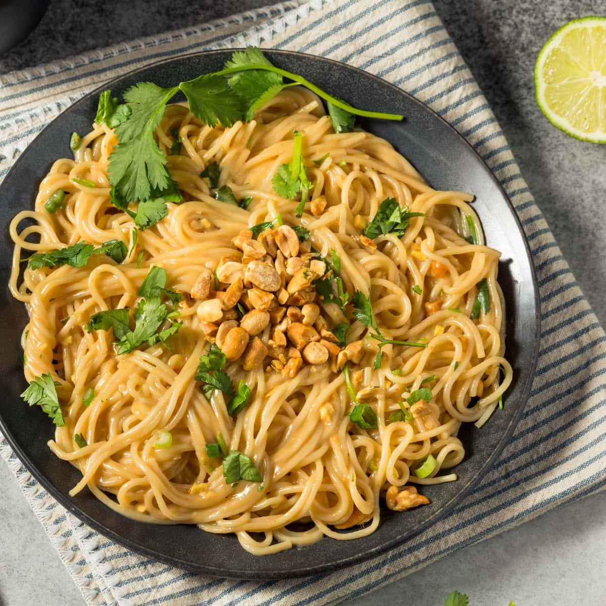 Noodle with peanut sauce served on a black plate garnished with chopped nuts and cilantro leaves.