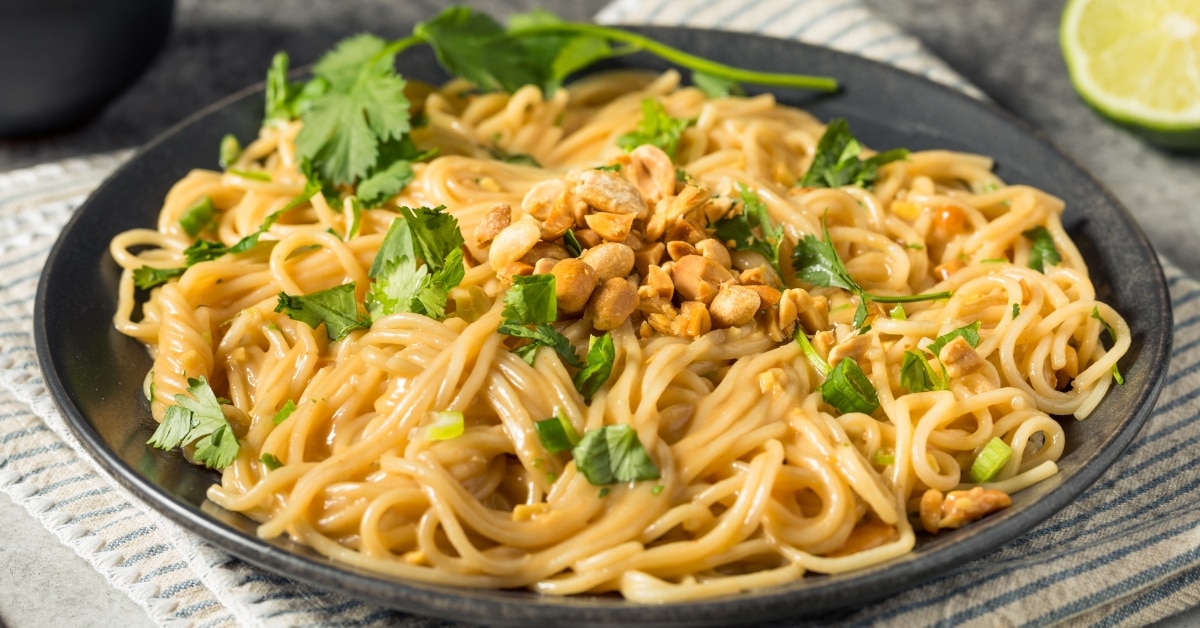 Peanut noodles on a black plate with chopped nuts and cilantro leaves garnish.