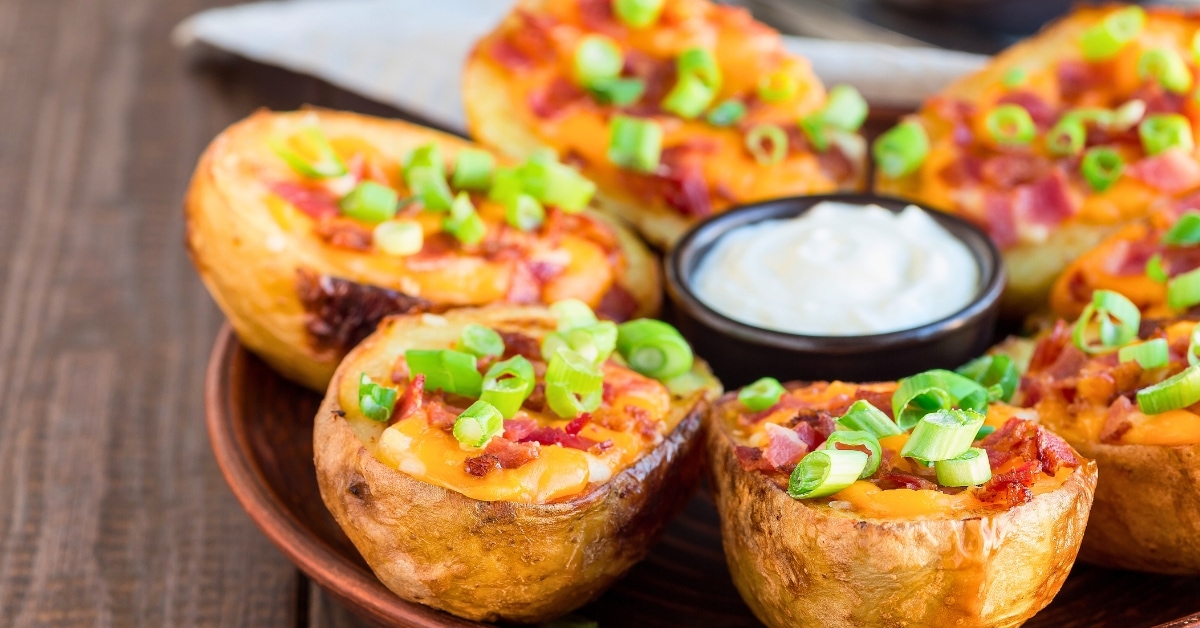 Baked potato skins with cheesy filling garnished with chopped onions with sour cream dip served on a wooden tray.