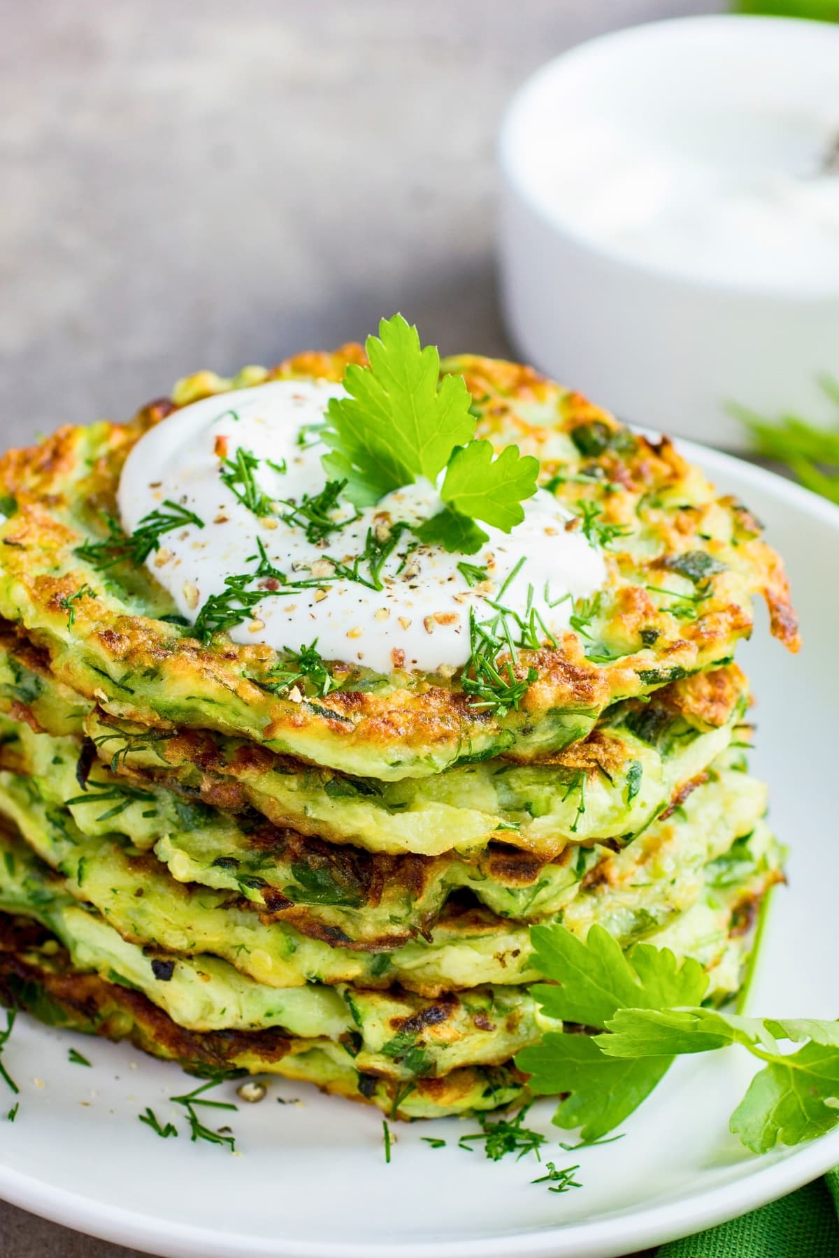 Stack of zucchini fritters with yogurt sauce and fresh dill garnish on top.