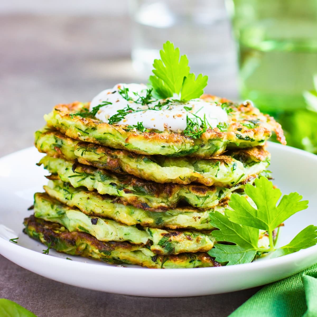 Stacks of zucchini fritters with yogurt on top garnished with dill and parsley leaves.
