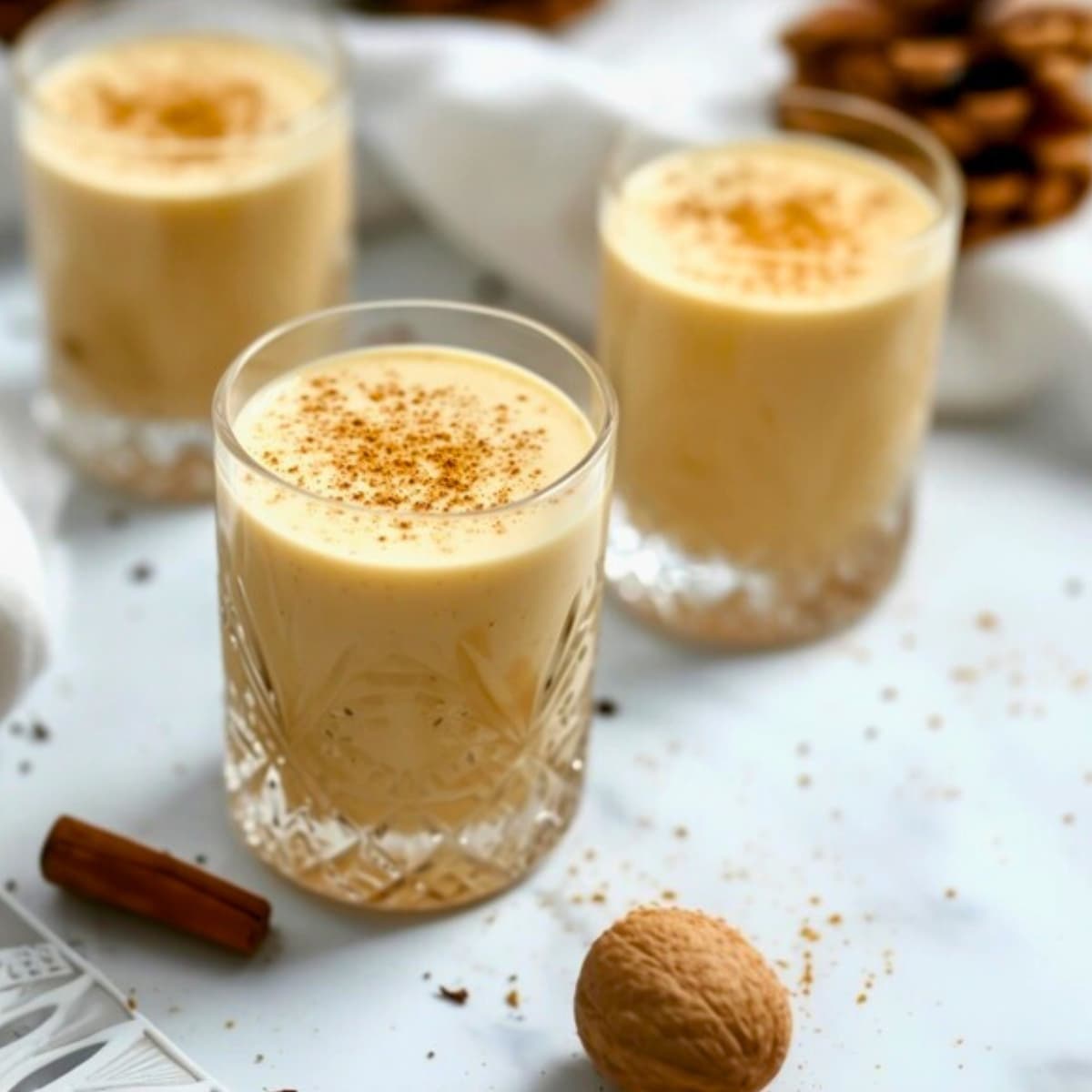 Three glasses of homemade eggnog on a white marble table garnished with grated nutmeg.