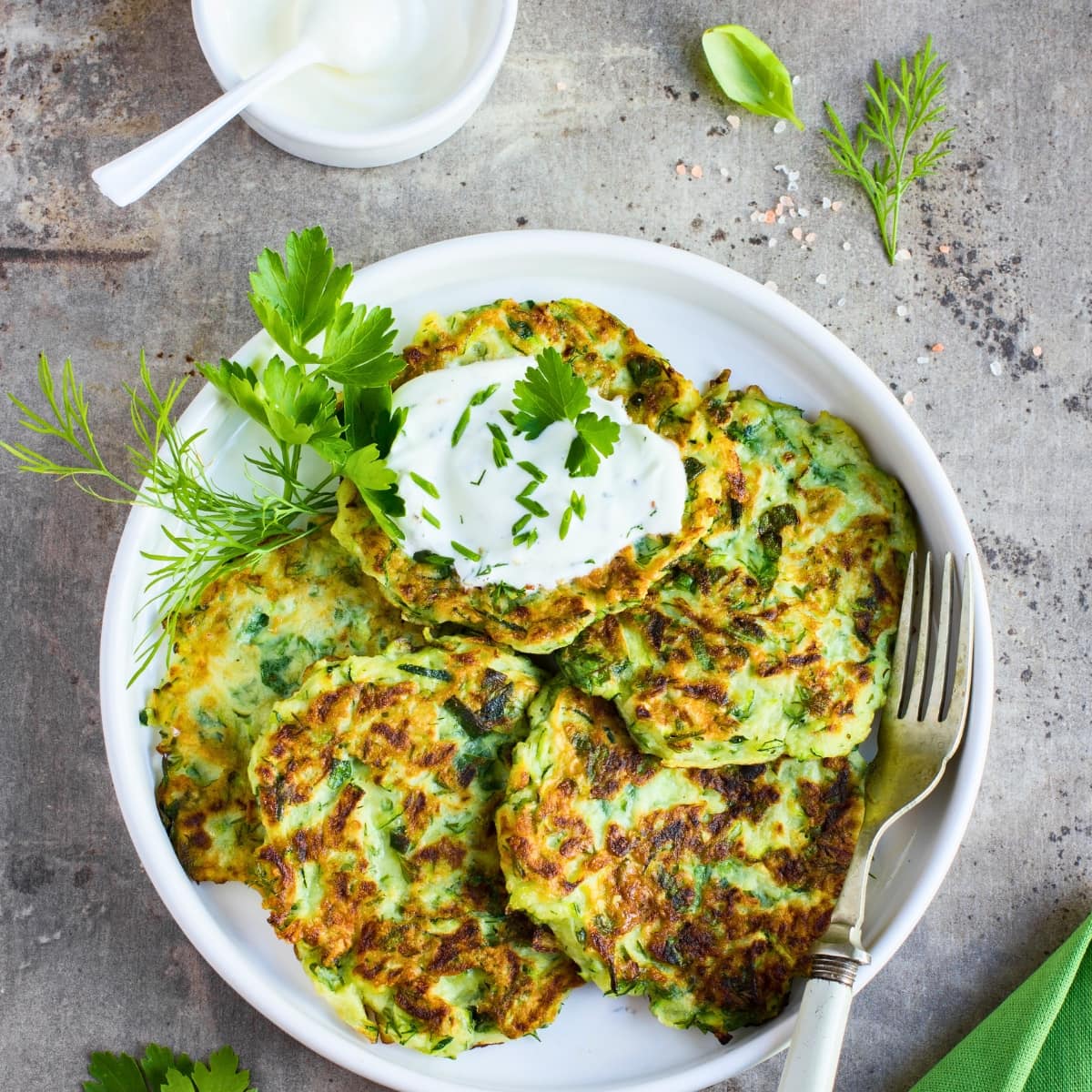 Zucchini fritters in a plate served with yogurt cream garnished with fresh dill and parsley leaves.