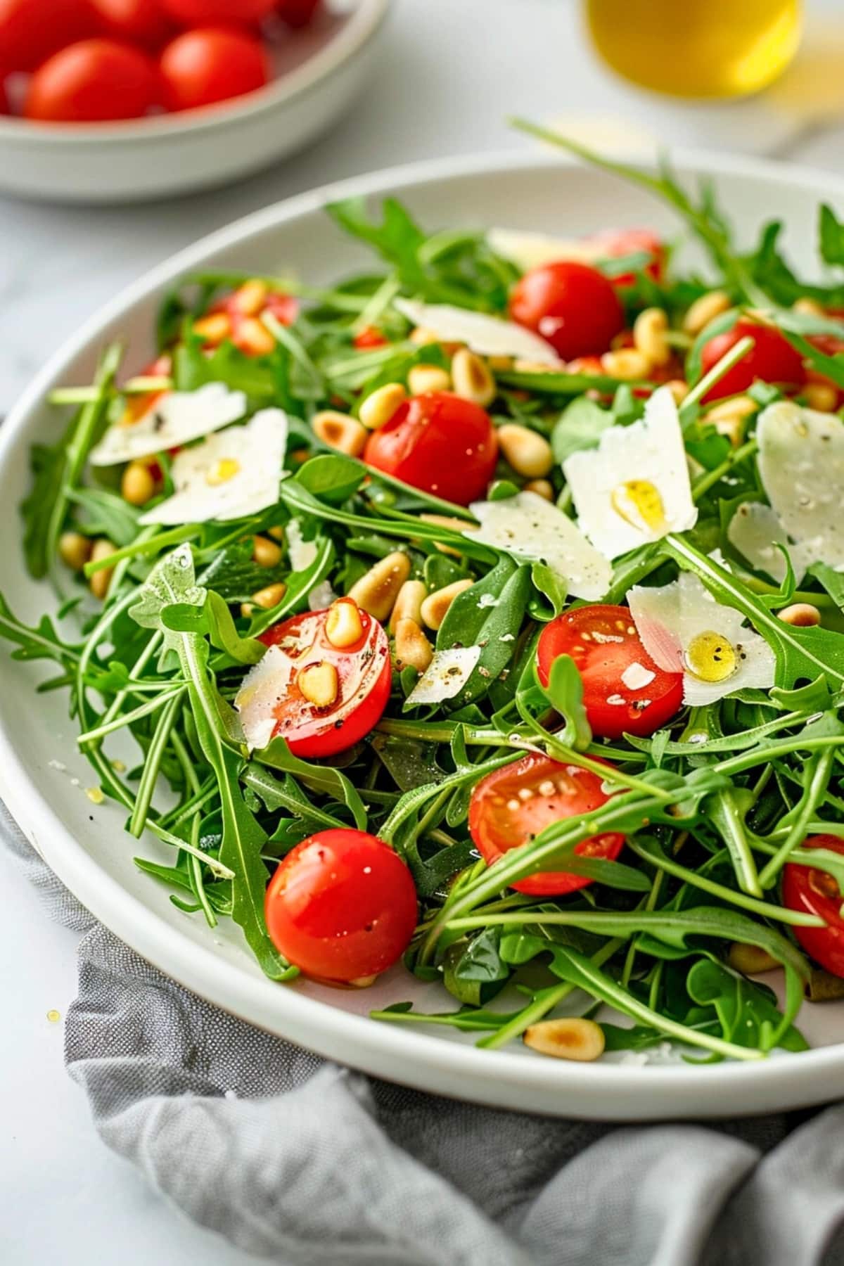 Arugula salad with cherry tomatoes, arugula and roasted pine nuts drizzled with lemon dressing garnished with shaved parmesan cheese on a white plate.