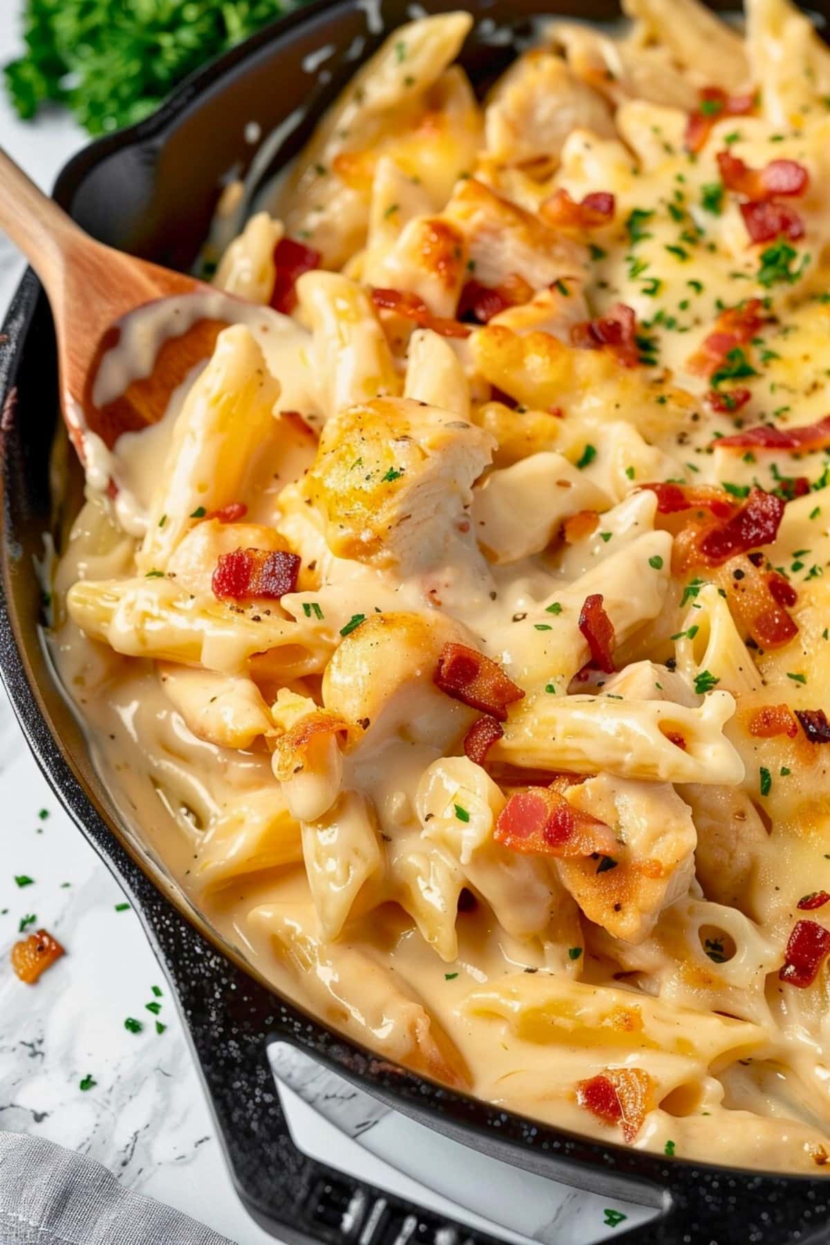 Penne pasta in cheesy and creamy sauce, bacon and chicken, tossed in a cast iron skillet with a wooden ladle.