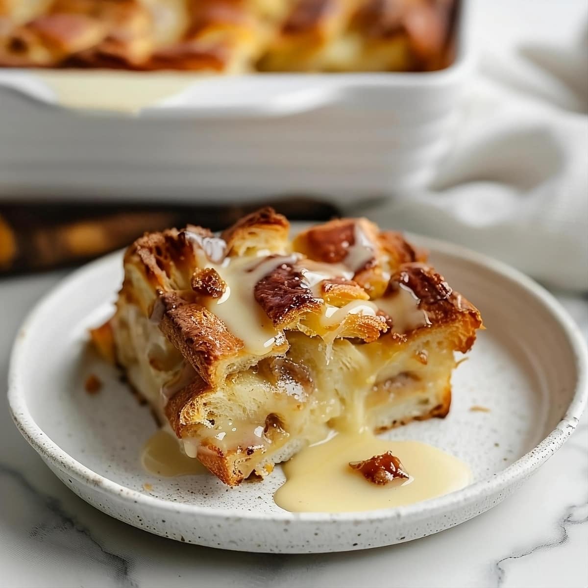 Slice of Croissant Bread Pudding on a plate dripping with vanilla sauce.