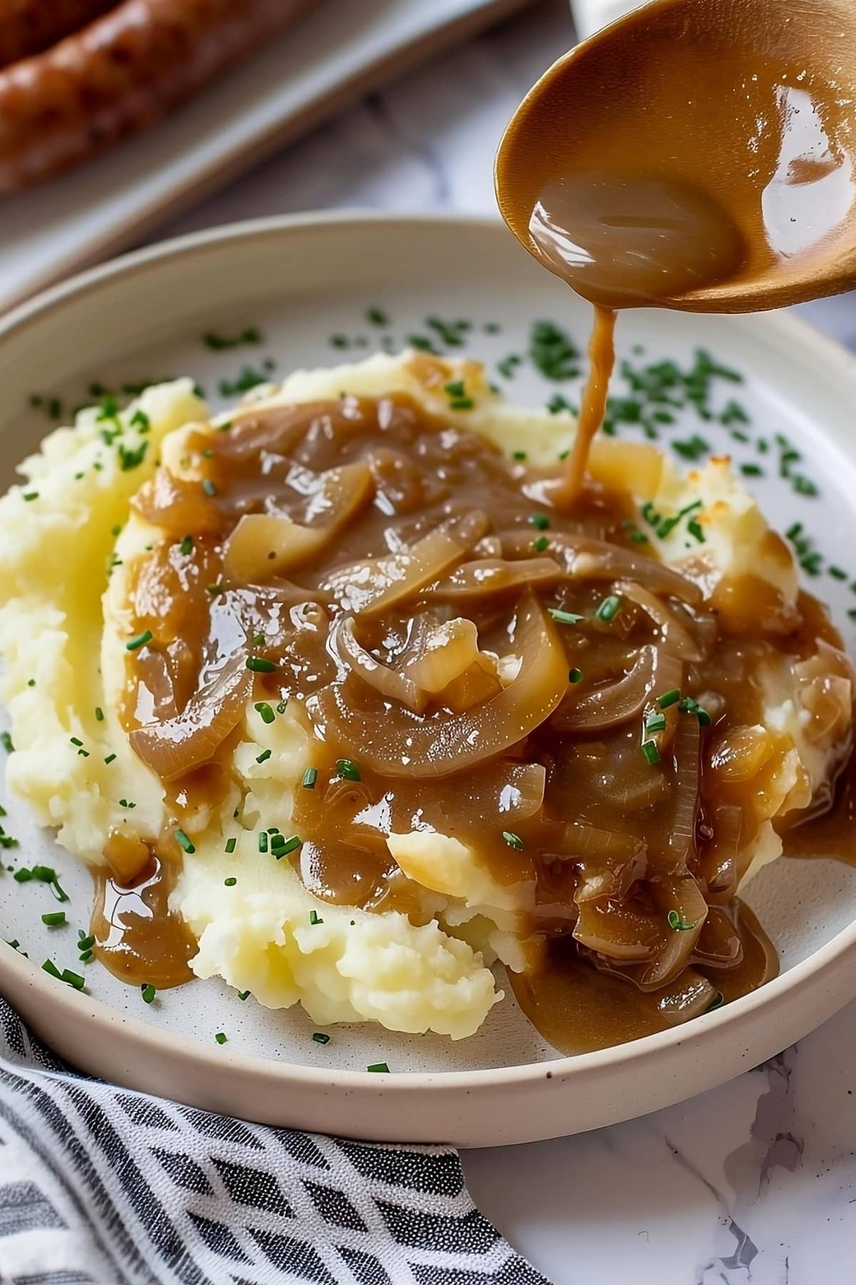 Mashed potato poured with onion gravy, sausage in plate in the background.