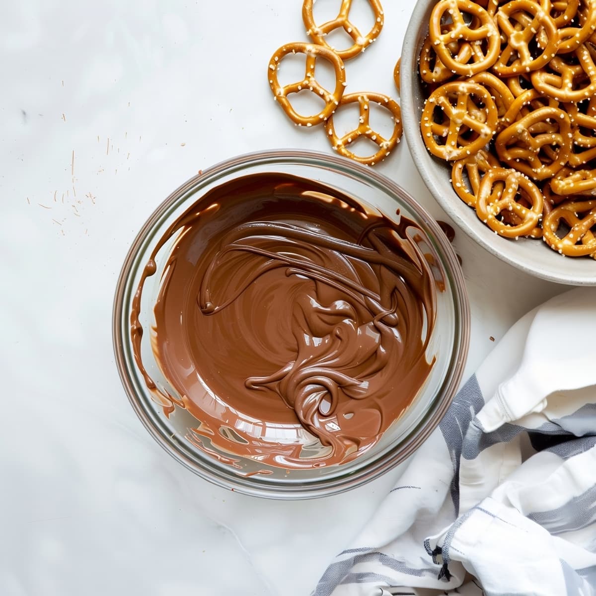A glass bowl of melted chocolate chips with pretzels on the side