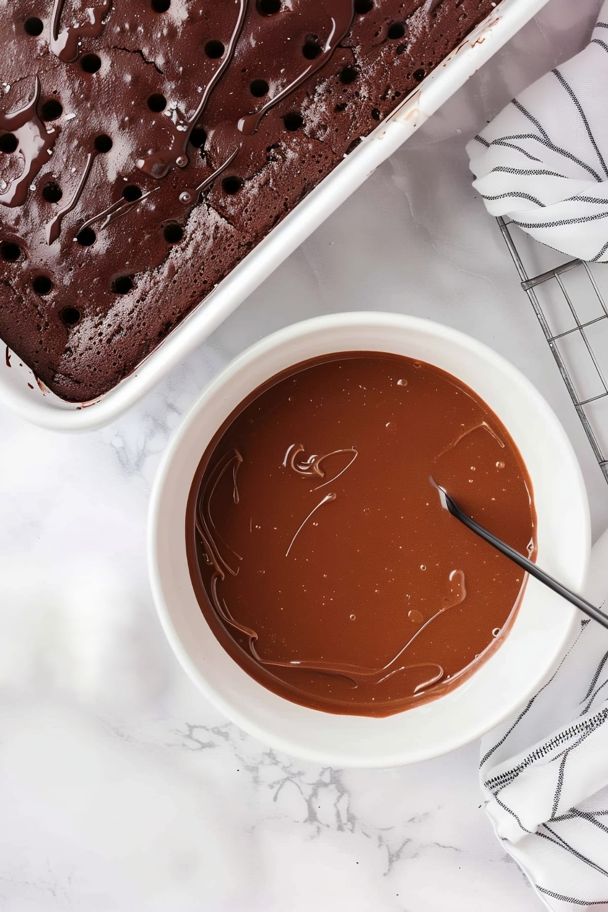 Overhead view of melted chocolate in a bowl with chocolate cake with holes on the side on a white marble table