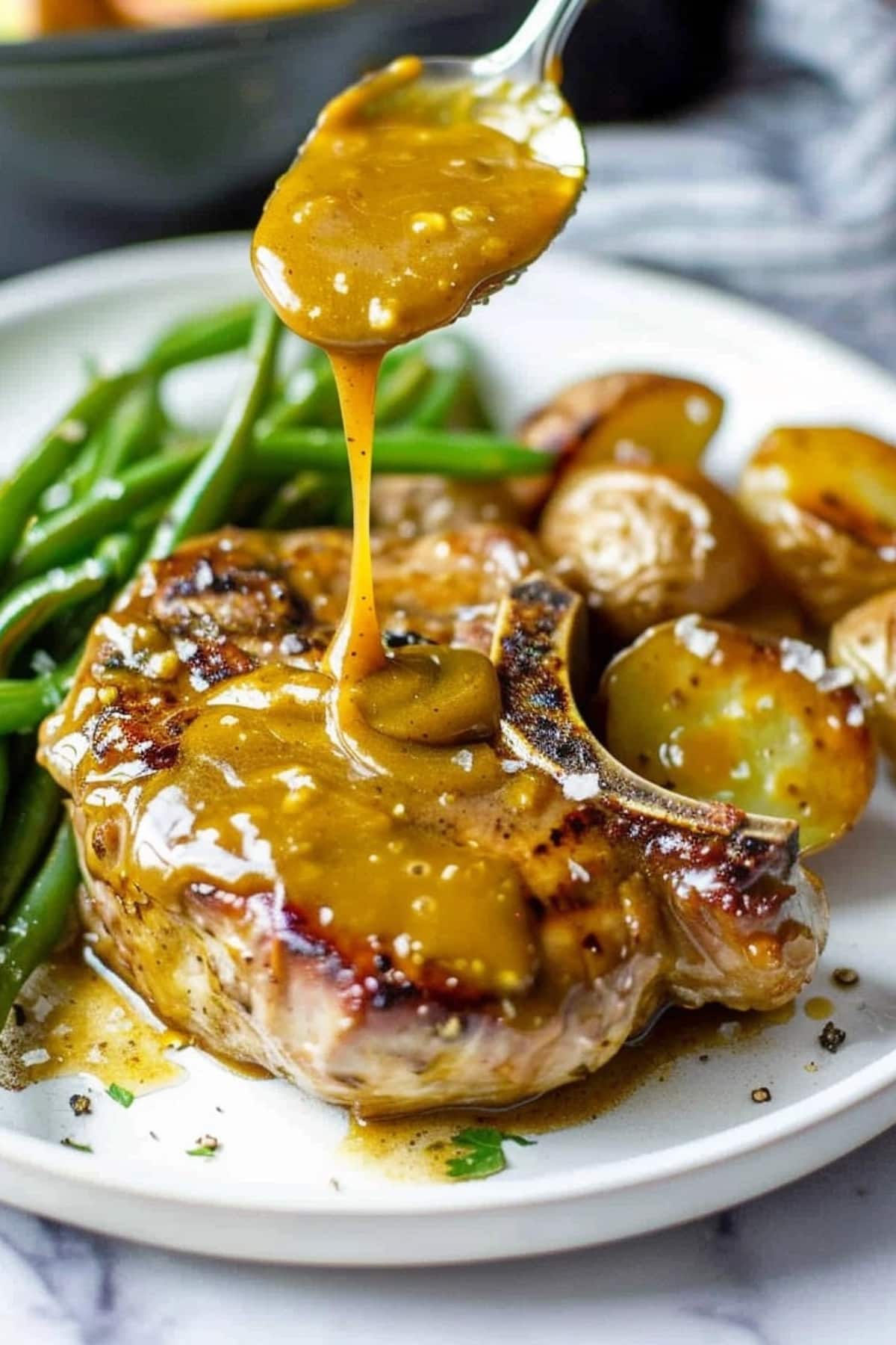Honey mustard sauce dripping on pork chop with beans and potatoes on the side.