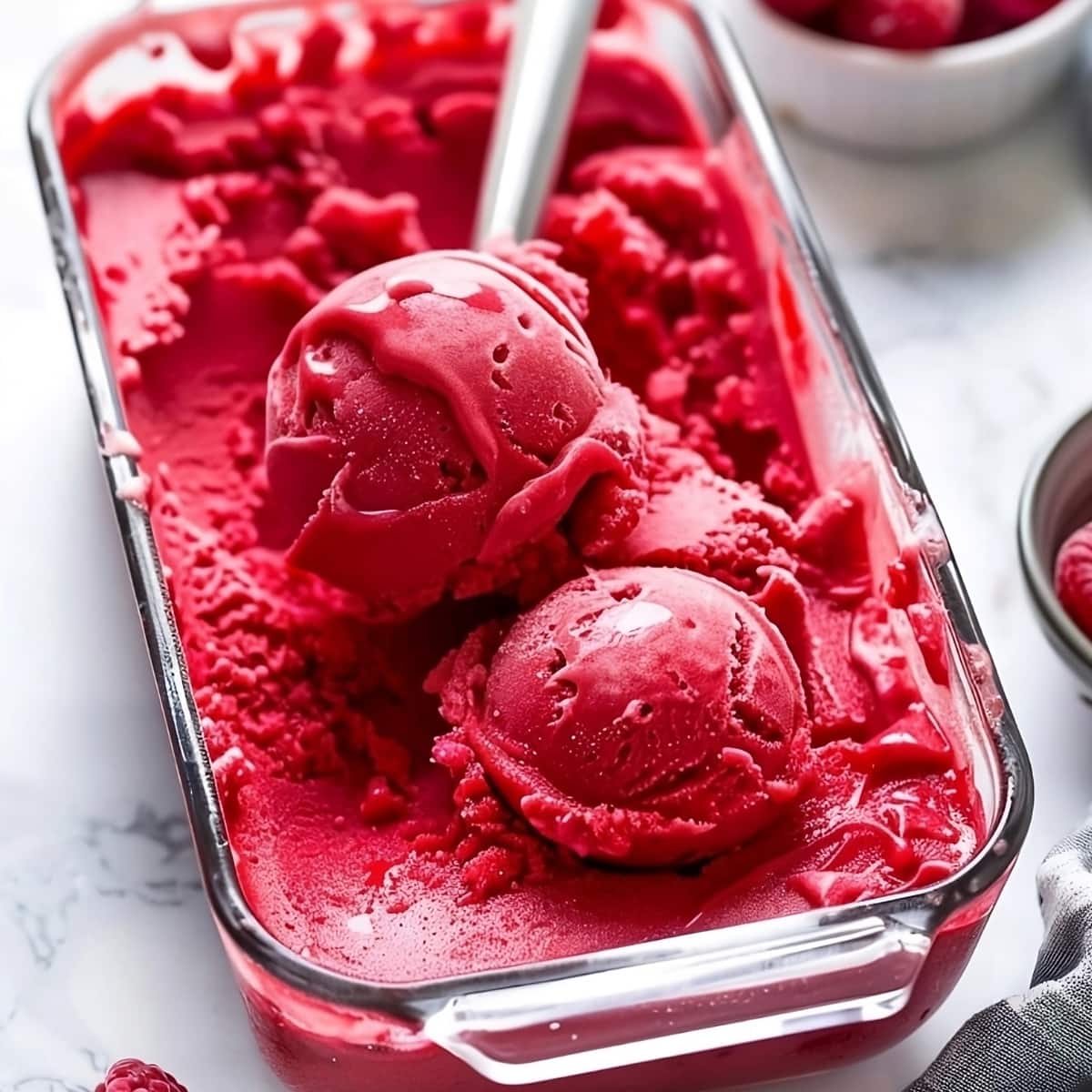 Raspberry sorbet in a glass dish with ice cream scooper.