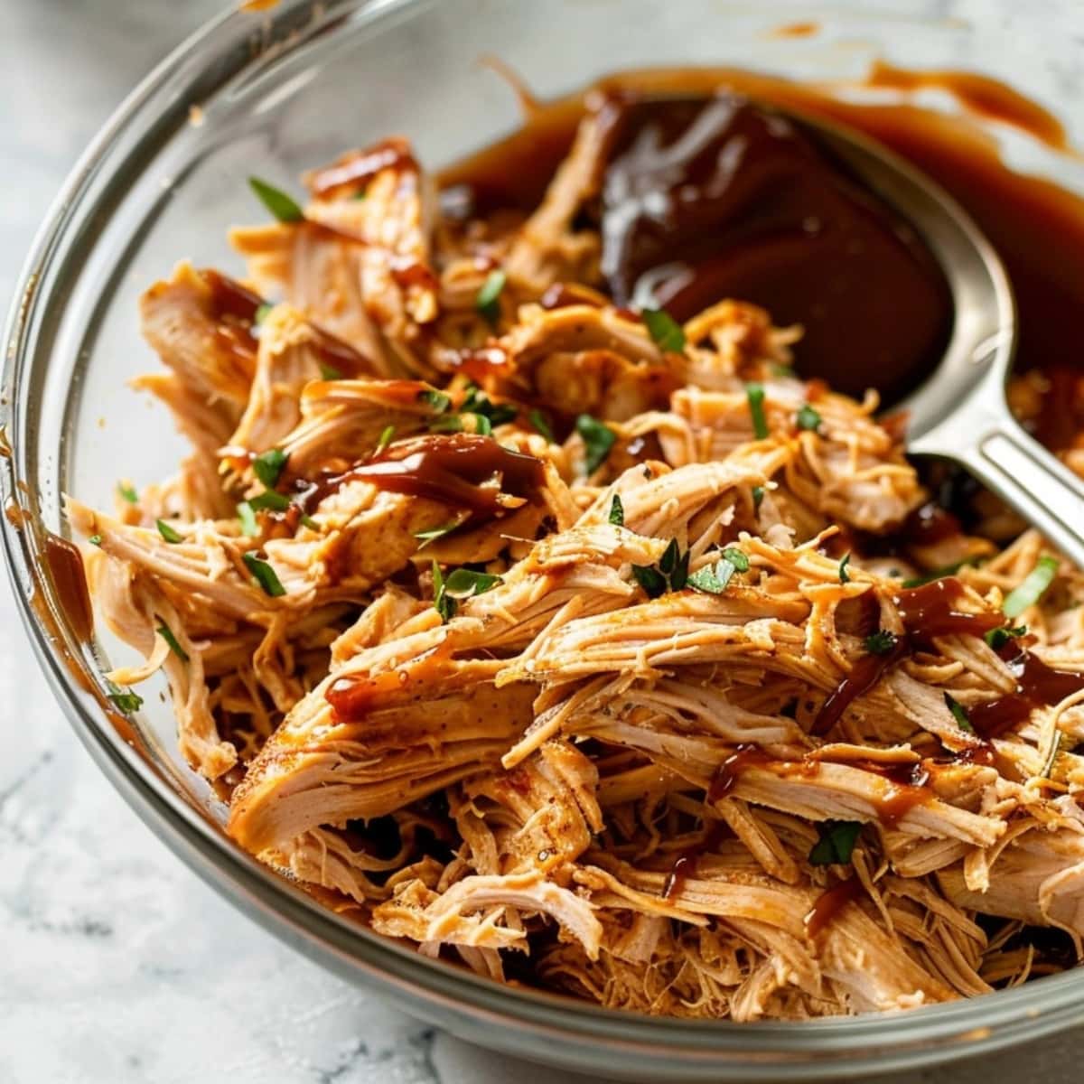 Shredded chicken with spoonful of bbq sauce on a clear glass bowl.