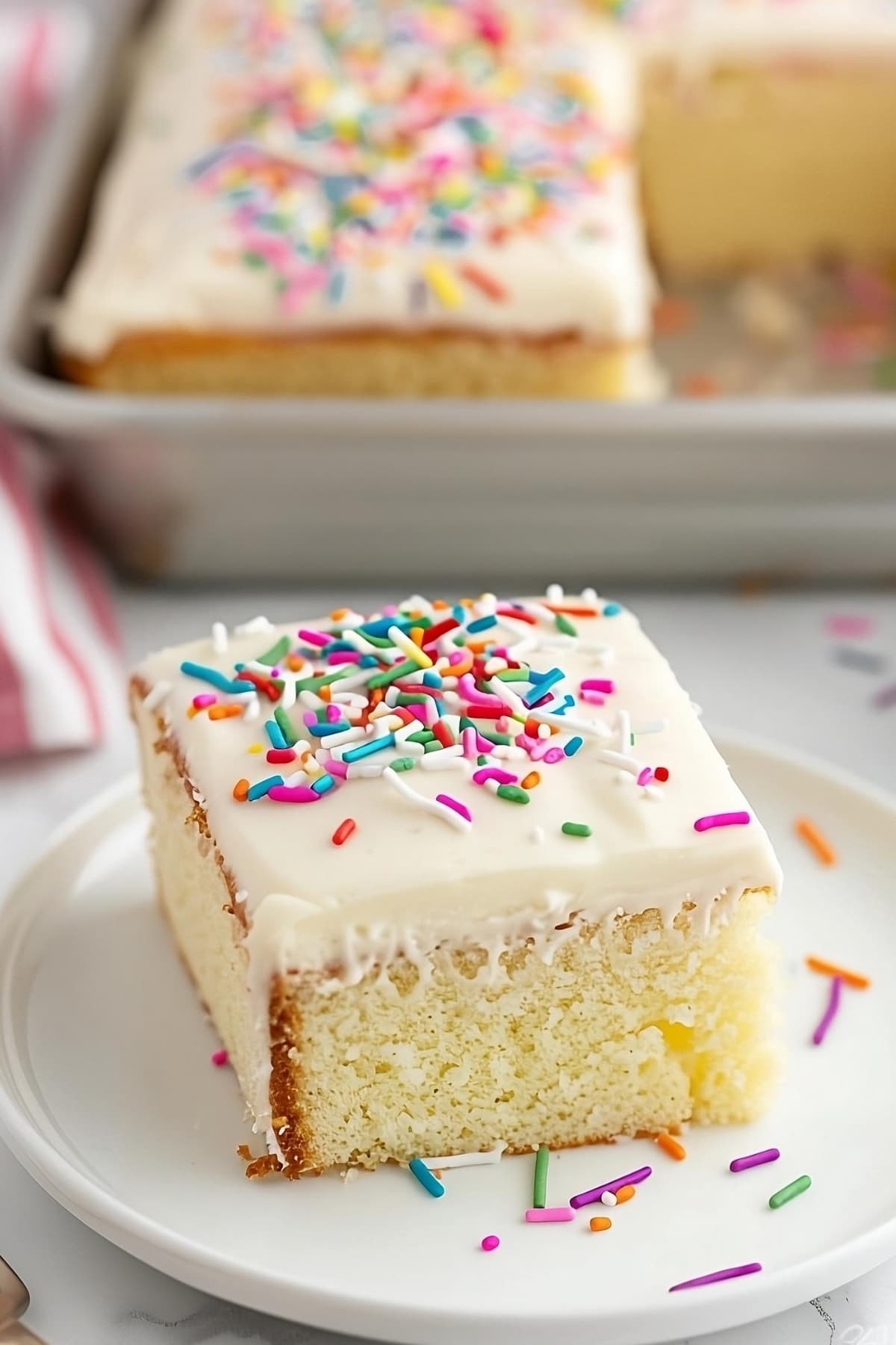 Square slice of vanilla sheet cake with rainbow sprinkles on a white plate.