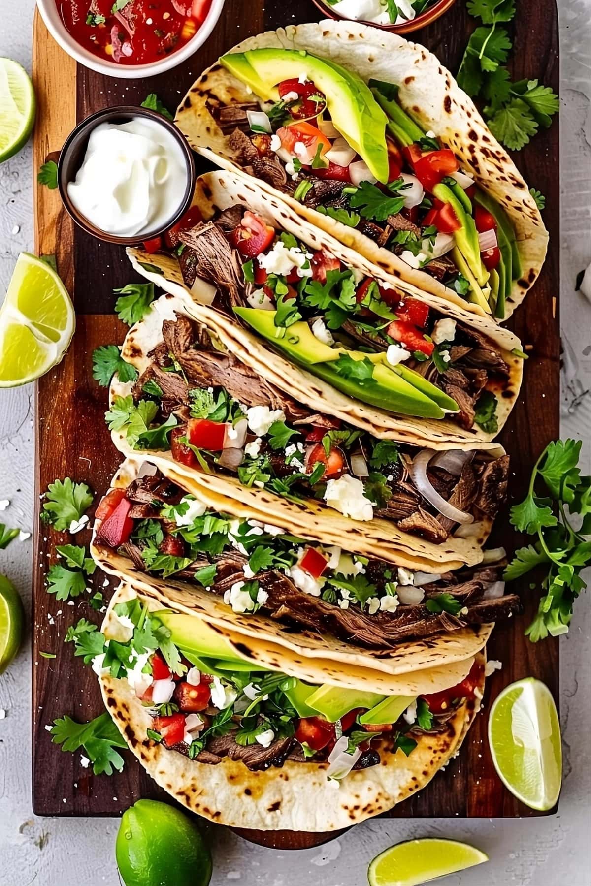 Tacos with shredded steak, onions and garlic, avocado, cilantro, sour cream, salsa, and queso fresco served on a wooden tray.