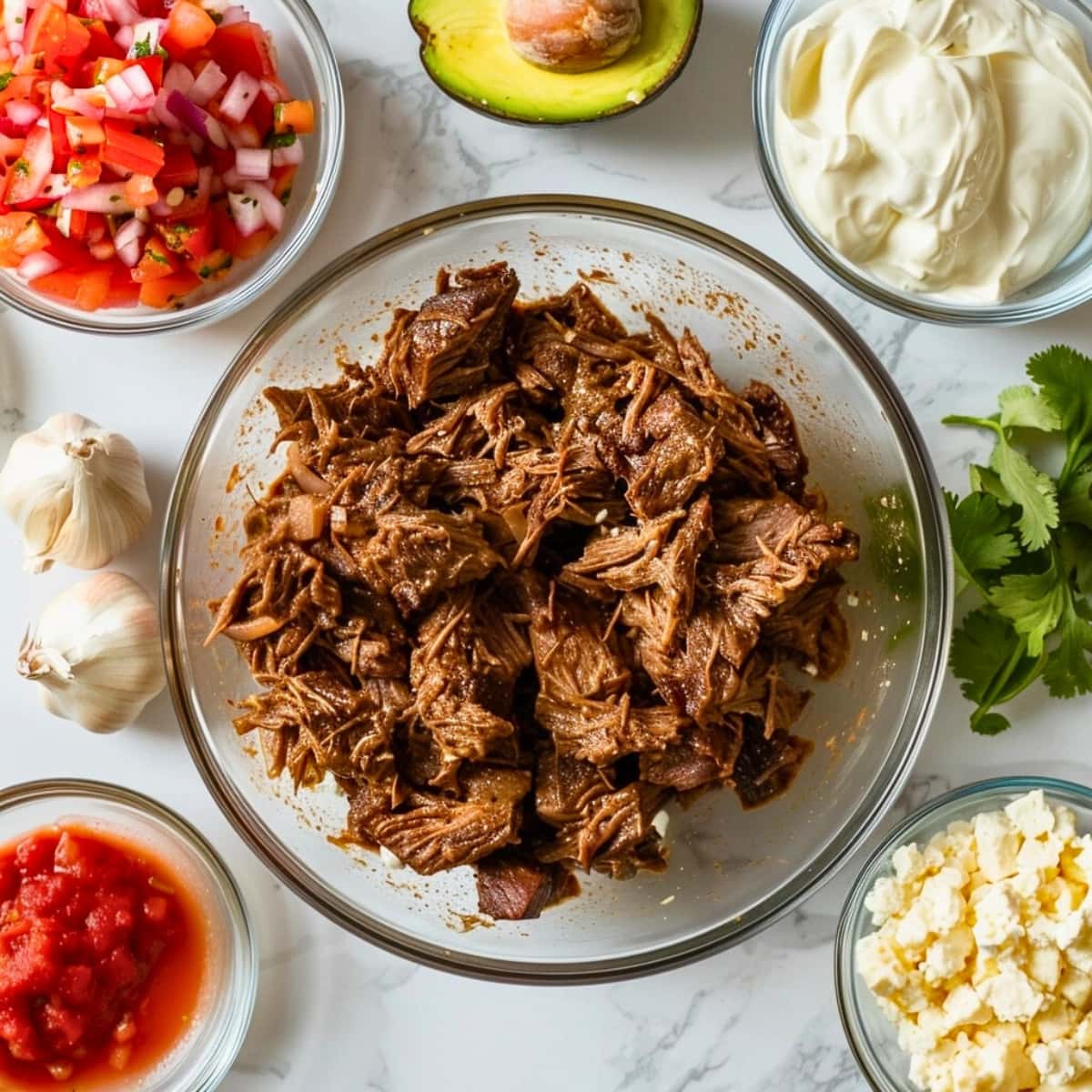 Shredded steak in a glass bowl, onions and garlic, avocado, cilantro, sour cream, salsa, and queso fresco in individual bowls on a white marble table top view.