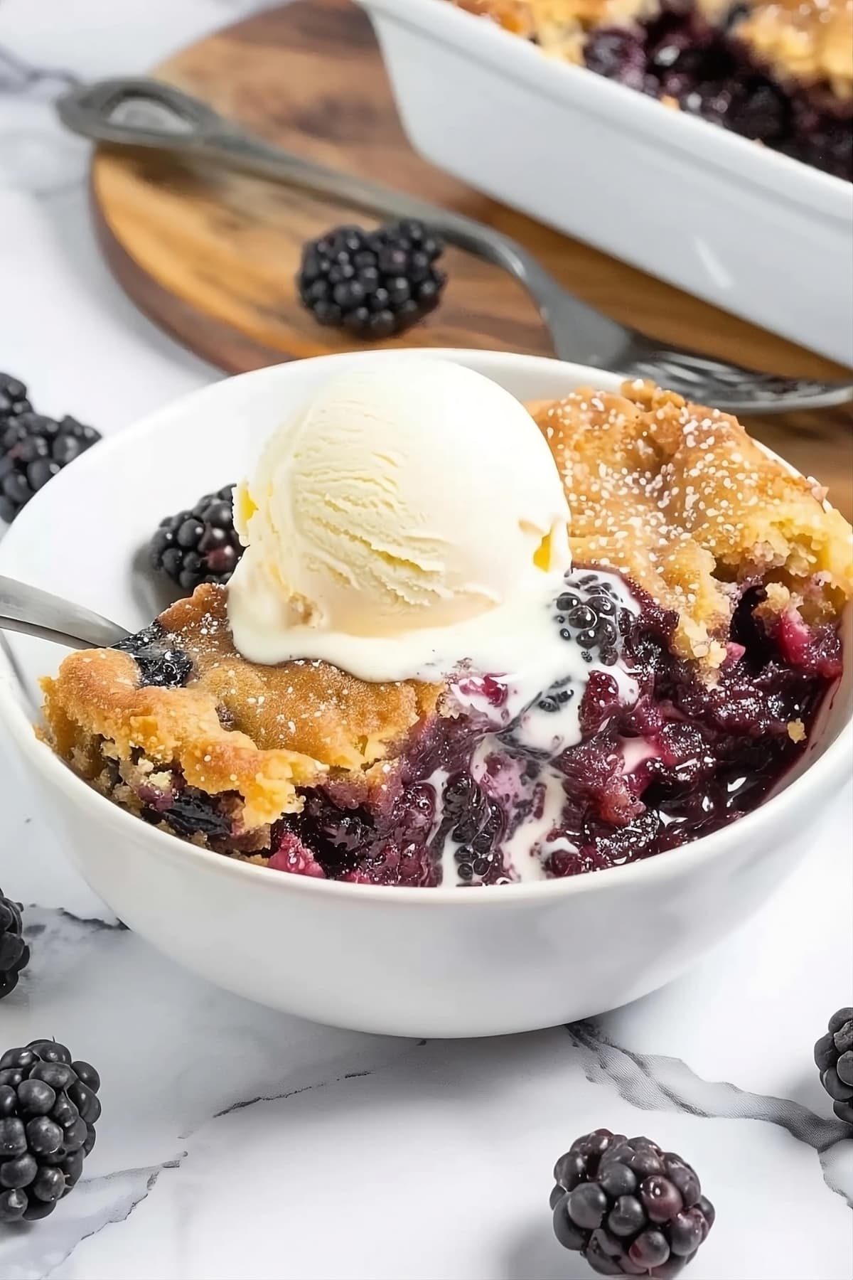 Blackberry dump cake in a white bowl with spoon garnished with vanilla ice cream scoop.