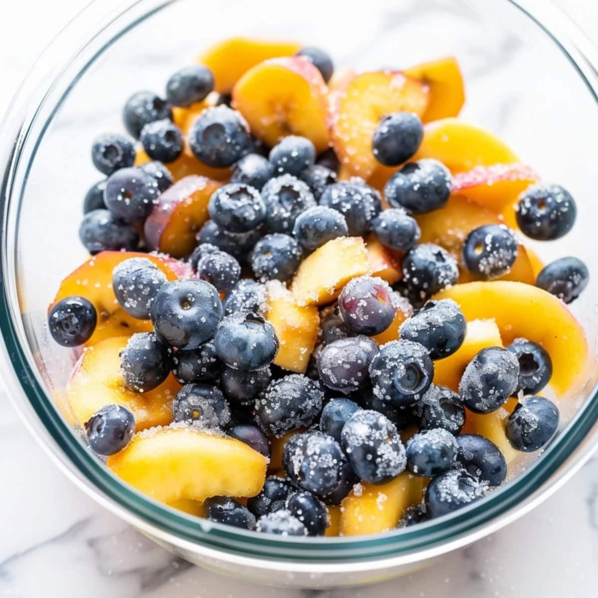 Sliced peaches and blueberries tossed in sugar in a glass bowl.