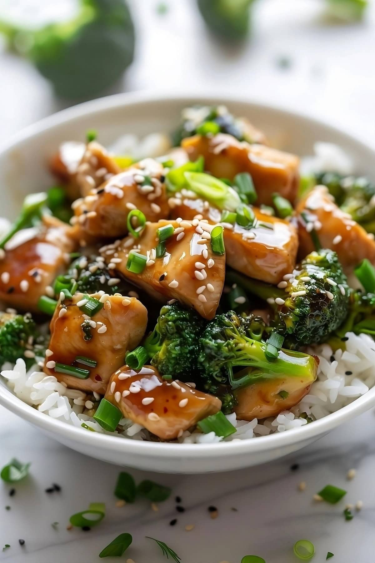 Bowl of saucy stir fried chicken and broccoli on top of white rice, garnished with spring onion and sesame seeds.
